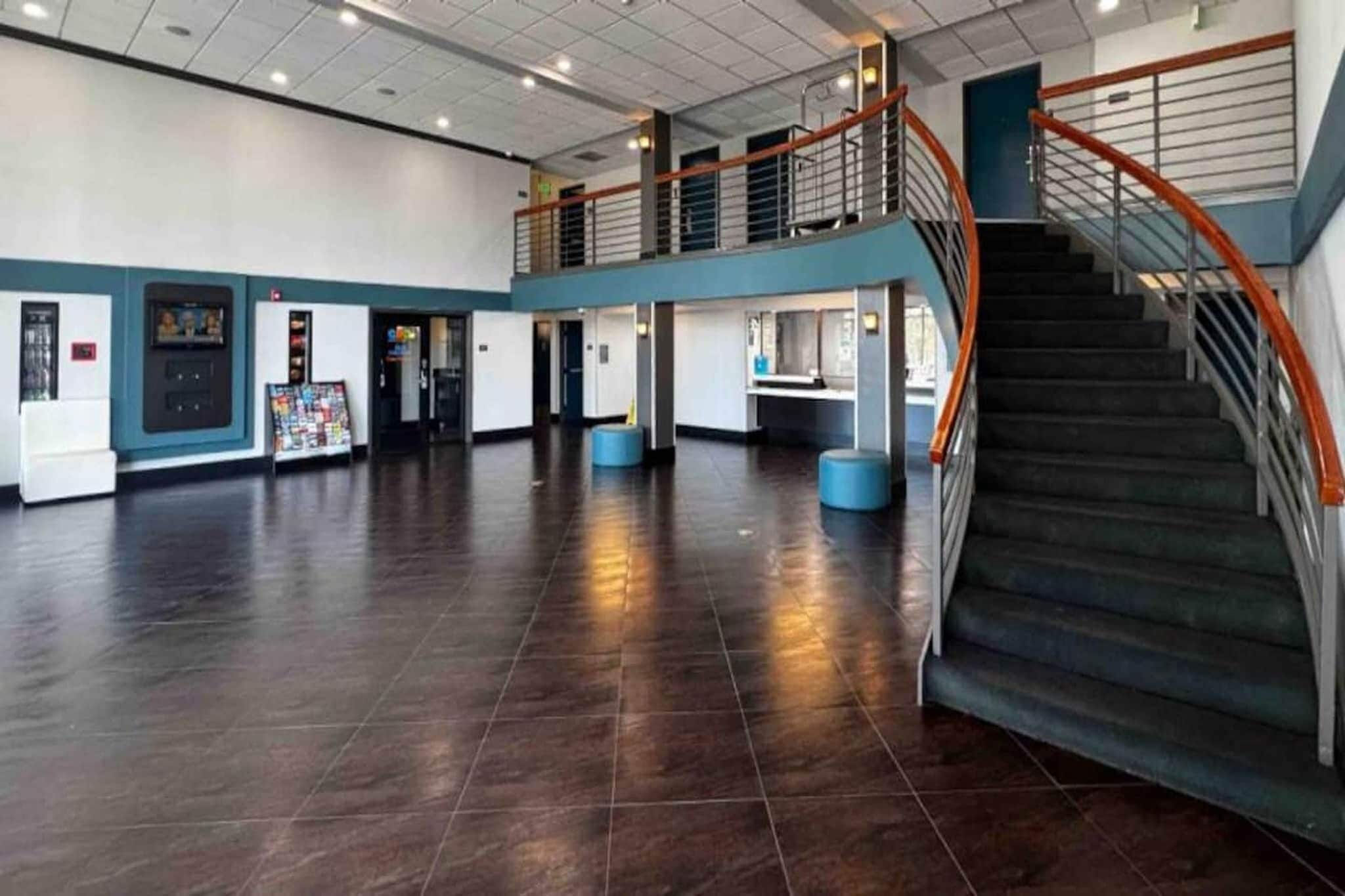A spacious lobby with a dark tiled floor, a staircase with orange railings, and a mezzanine level with a railing.