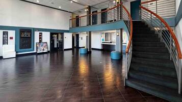 A spacious lobby with a dark tiled floor, a staircase with orange railings, and a mezzanine level with a railing.
