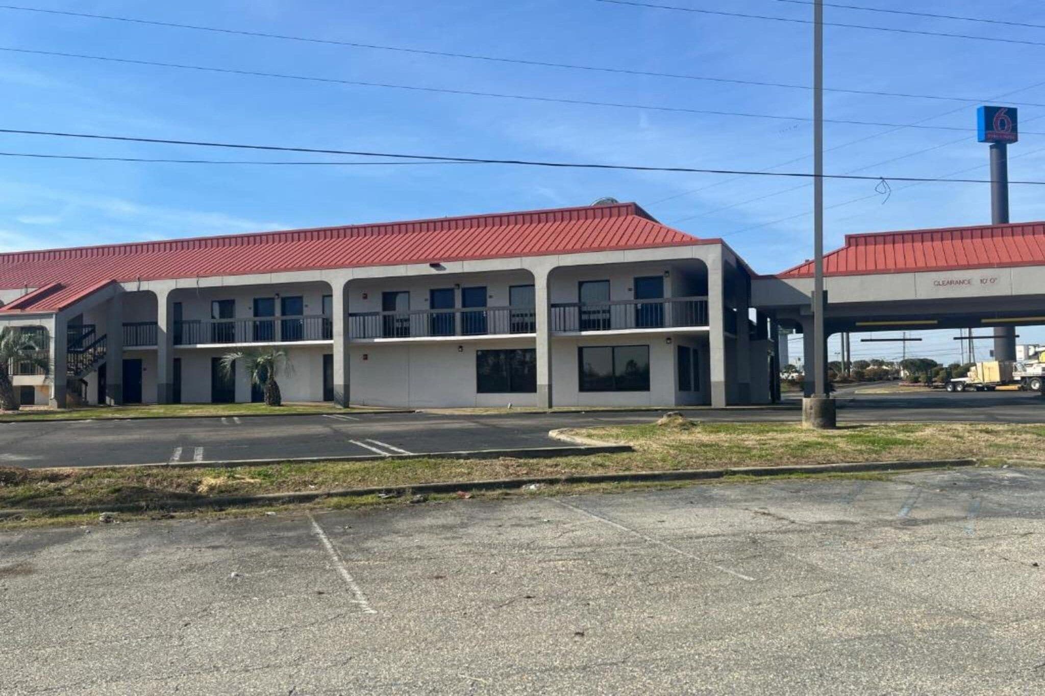 A two-story motel with a red roof, a parking lot, and a gas station in the background.