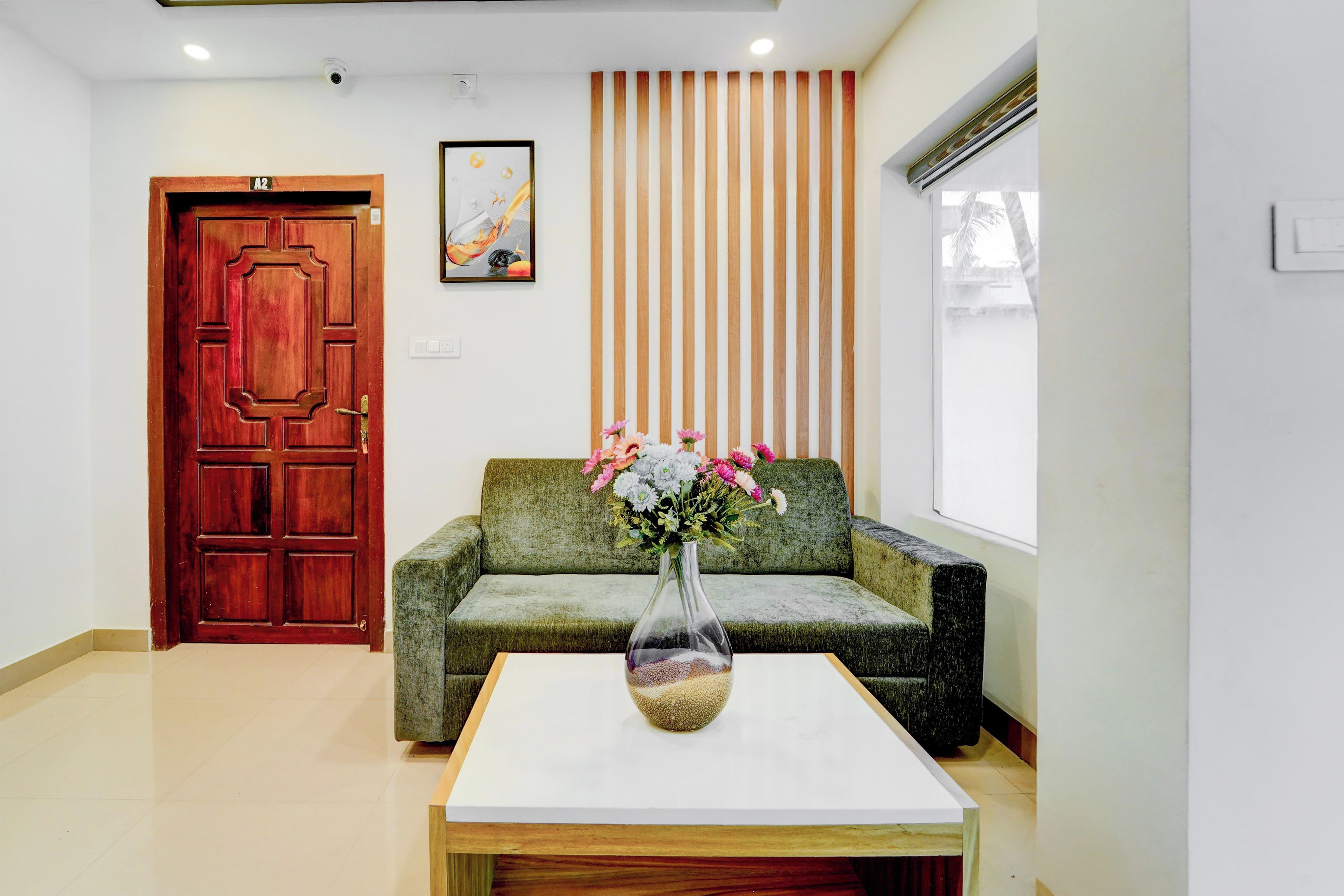 Un área de recepción con una puerta de madera, un sofá verde, una mesa blanca con un jarrón de flores y un panel de madera vertical en la pared.