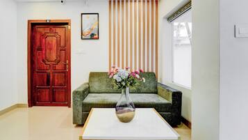 Un área de recepción con una puerta de madera, un sofá verde, una mesa blanca con un jarrón de flores y un panel de madera vertical en la pared.