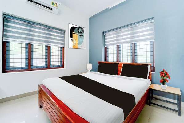 A modern bedroom with a large bed, two windows with blinds, a wall-mounted air conditioner, and a framed abstract painting.