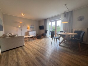 A modern living room with a sofa, a dining table with chairs, a TV mounted on the wall, and a large window with curtains. - Jacobihof - Finn - "Finn," a 3-room apartment near the beach in a quiet cul-de-sac (Dahme)