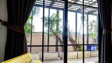 Una silla amarilla con un cojín de hoja verde, una cortina negra y vistas a través de amplias ventanas de un espacio exterior con estructuras de concreto y vegetación.