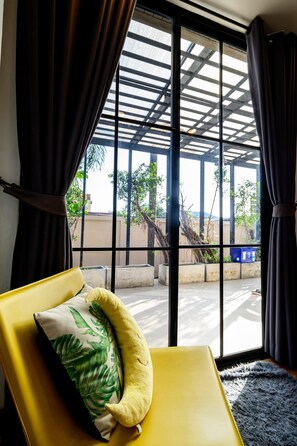 A yellow chair with a green leaf pillow, a black curtain, and a view through large windows to an outdoor area with concrete structures and greenery. - Modern Spacious 3 beds Pool Villa BA1 (Pattaya)
