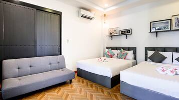Habitación de hotel con sofá, dos camas, piso con estampado de espiga, unidad de aire acondicionado y obras de arte colgadas en la pared.