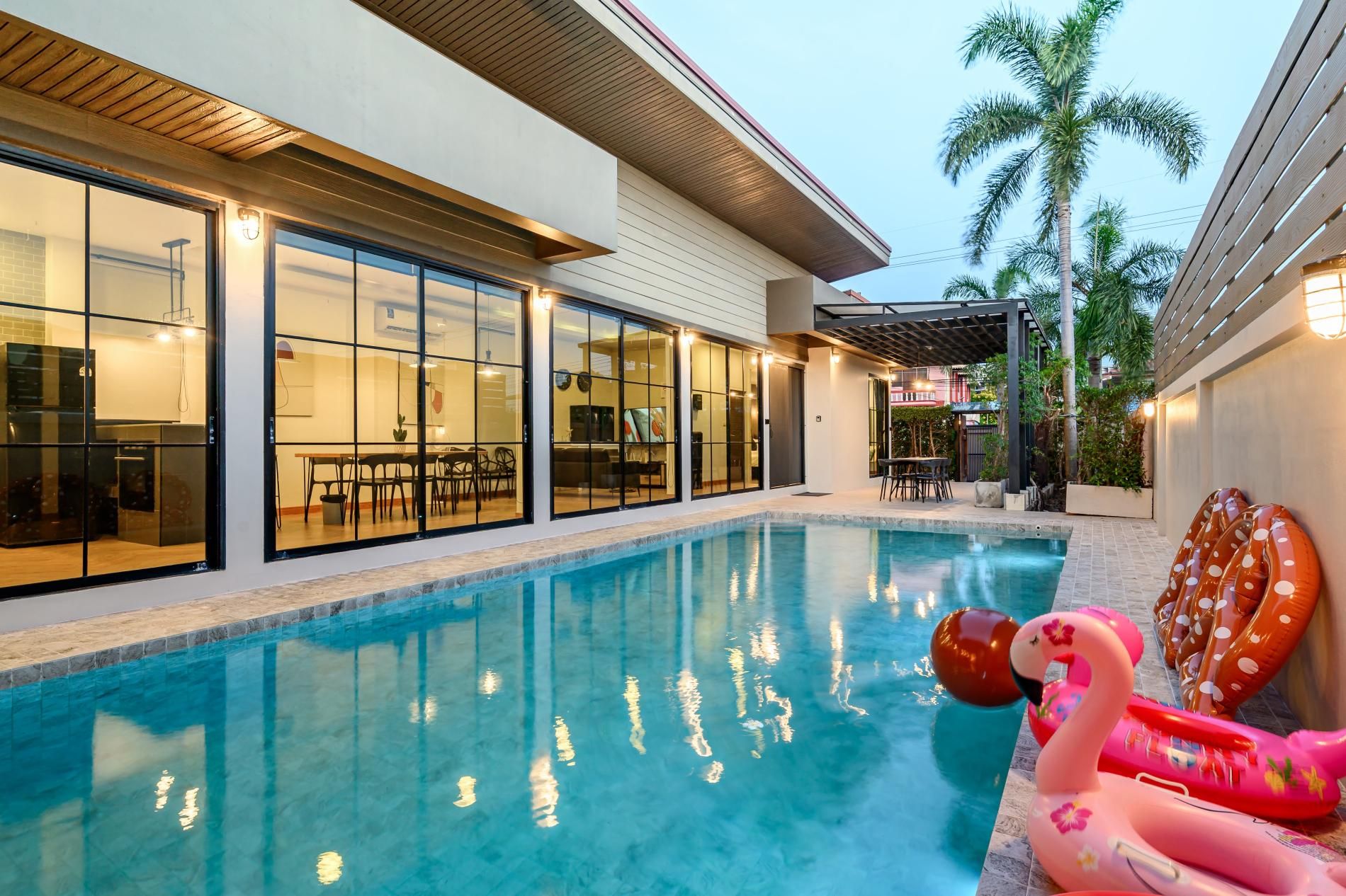 Una piscina con agua azul cristalina, rodeada por una casa moderna con grandes ventanales y un porche cubierto.