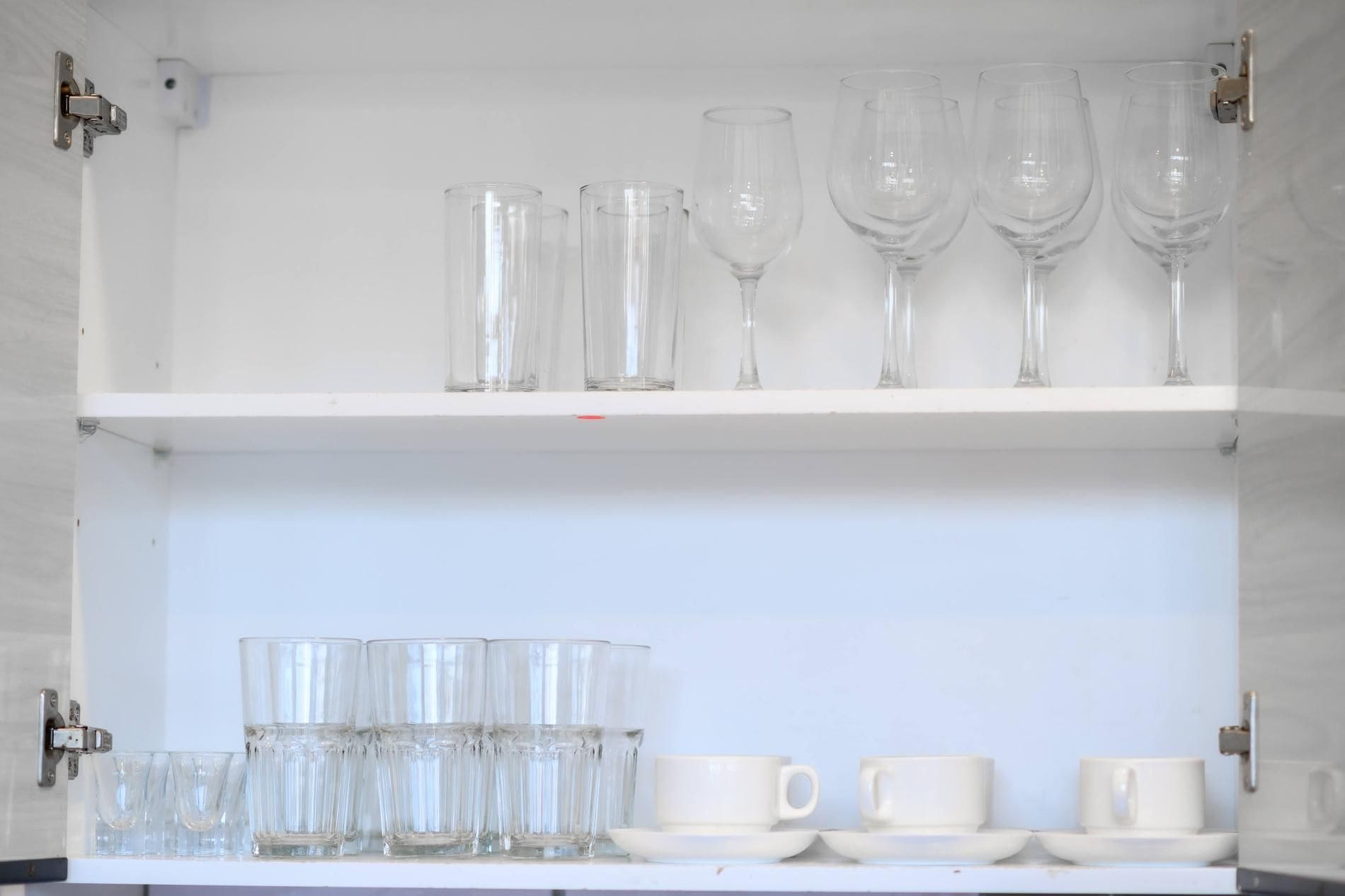 A white cabinet with two shelves containing clear glasses and white cups.