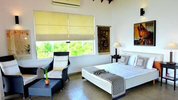 A bedroom with a bed, two chairs, a small table, a nightstand, a lamp, and a window with a view of greenery.