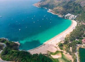 An aerial view of a coastal area with a beach, boats, and a residential area with buildings. - Sunrise Studio, Nai Harn, Desk, Rooftop Pool (Tambon Rawai)
