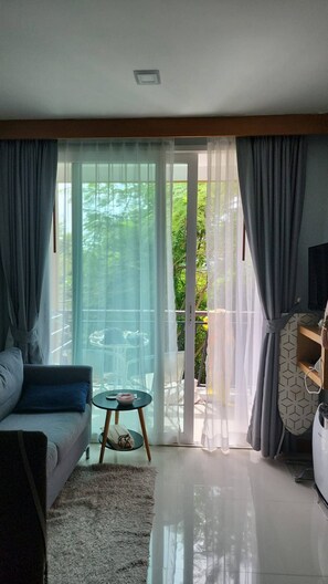 A modern living room with a sofa, a small round table, and a sliding glass door leading to a balcony. - Quiet & Cozy 1BR, Pool, Rawai, Walk to Gyms (Rawai)