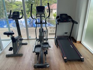 A home gym with cardio equipment including a treadmill, elliptical, and stationary bike, with a view of a swimming pool outside. - Quiet & Cozy 1BR, Pool, Rawai, Walk to Gyms (Rawai)