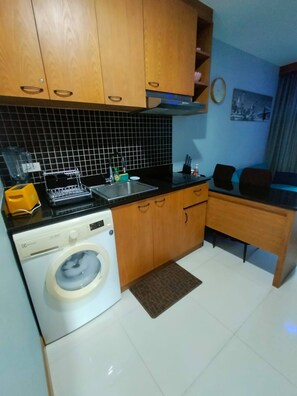 A compact kitchen with wooden cabinets, a washing machine, a sink, and a dining area with a chair. - Quiet & Cozy 1BR, Pool, Rawai, Walk to Gyms (Rawai)