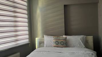A hotel room with a bed, a window with blinds, a radiator, and a light fixture.