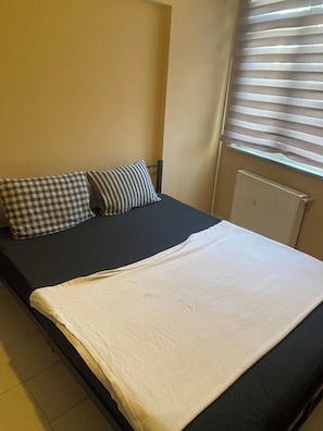 A single bed with a checkered pillow and a striped pillow, a white blanket, and a window with blinds.