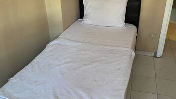 A single bed with a white bedspread and a black headboard in a room with beige walls and tiled flooring.