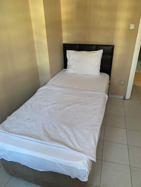 Ein Einzelbett mit weißer Bettdecke und einem schwarzen Kopfteil in einem Zimmer mit beige gestrichenen Wänden und Fliesenboden.