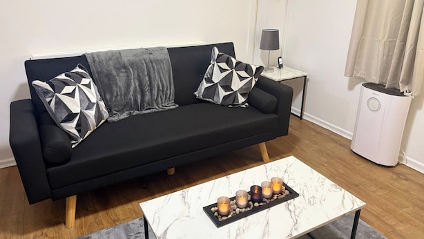 Um sofá preto com almofadas estampadas em padrão geométrico, uma mesa de centro de mármore com velas, uma pequena mesinha de cabeceira com abajur e um radiador branco.