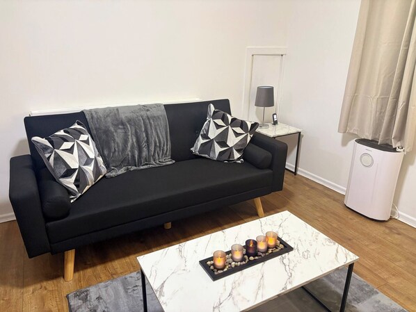 A black sofa with geometric patterned pillows, a marble coffee table with candles, a small bedside table with a lamp, and a white radiator. - Luxurious 3-Bedroom Apartment in Swansea City Centre (Swansea)
