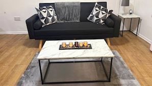 Uma sala de estar moderna com um sofá preto, duas almofadas decorativas estampadas, uma mesa de centro de mármore com velas e uma pequena mesa lateral com uma luminária.