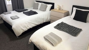 Quarto de hotel com duas camas, cada uma com edredom branco e cobertores cinzentos, uma mesa de cabeceira com abajur e uma janela com cortinas.