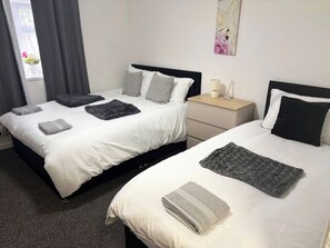 A hotel room with two beds, each with a white duvet and grey blankets, a bedside table with a lamp, and a window with curtains. - Luxurious 3-Bedroom Apartment in Swansea City Centre (Swansea)