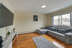 A modern living room with a sectional sofa, a flat-screen TV, a potted plant, and a window with a view of buildings.