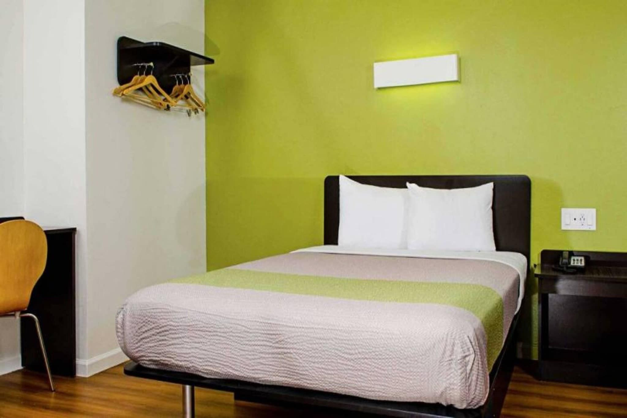 Habitación de hotel con cama, una silla, una mesita de noche y un estante con perchero.