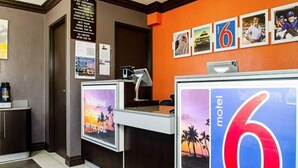 Mostrador de recepción de motel con un cartel grande con el número '6', una computadora y un cartel con un atardecer y palmeras.