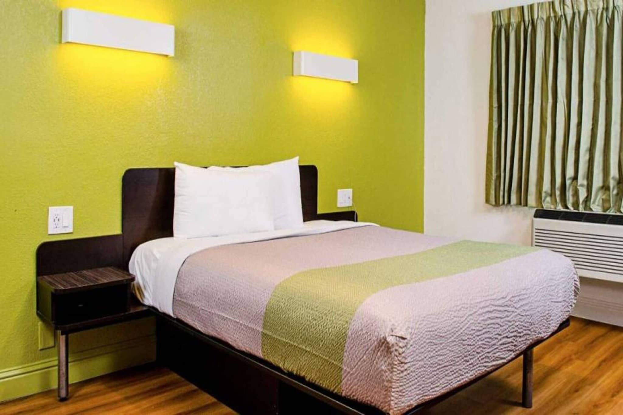 Habitación de hotel con cama, mesitas de noche, luz empotrada en la pared, ventana con cortinas y un calentador de pared.