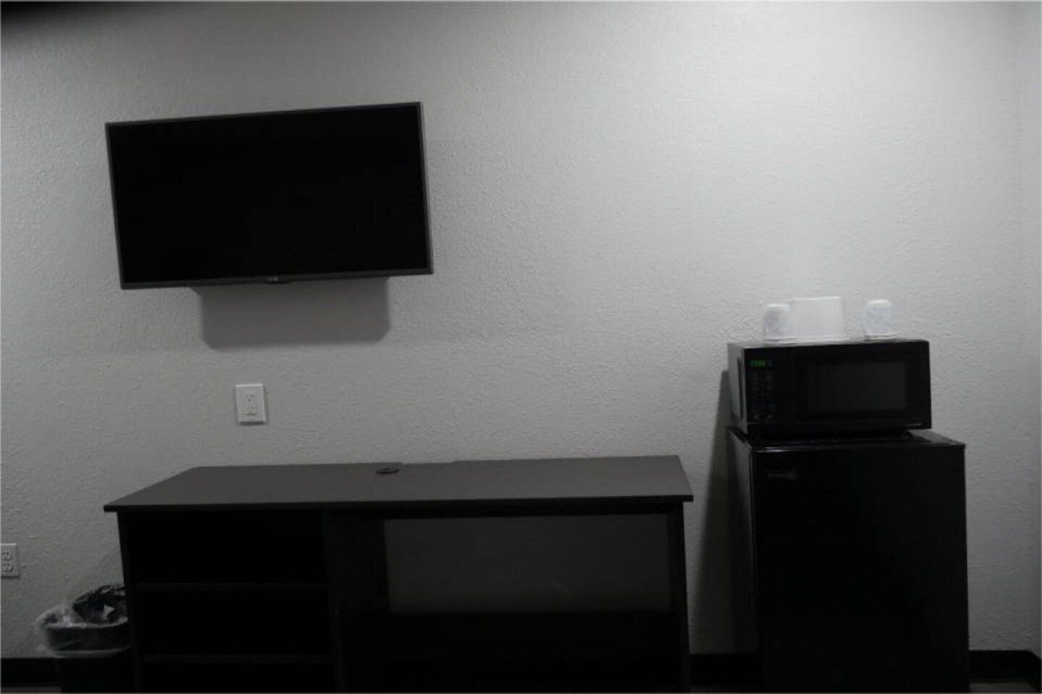 Une chambre dotée d’un téléviseur à écran plat fixé au mur, d’un petit réfrigérateur noir sur lequel est posé un four à micro-ondes, et d’un bureau noir avec une poubelle.