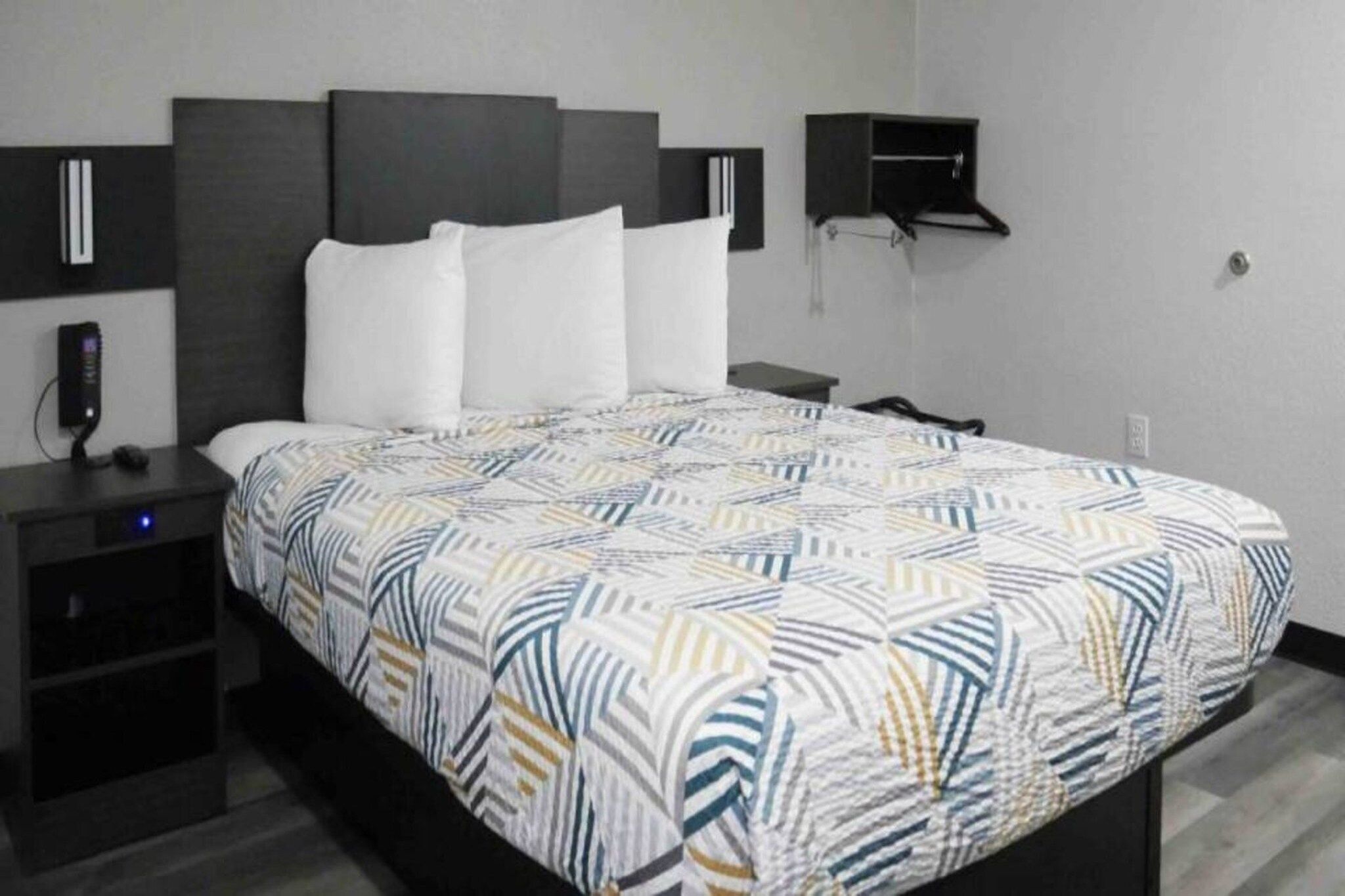 A hotel room with a bed, a bedside table, a nightstand, a wall-mounted shelf, and a power outlet.