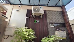 Une entrée d’immeuble équipée d’une caméra de surveillance, d’un climatiseur, d’une enseigne indiquant l’adresse et d’une petite table sur laquelle est posé un objet décoratif.