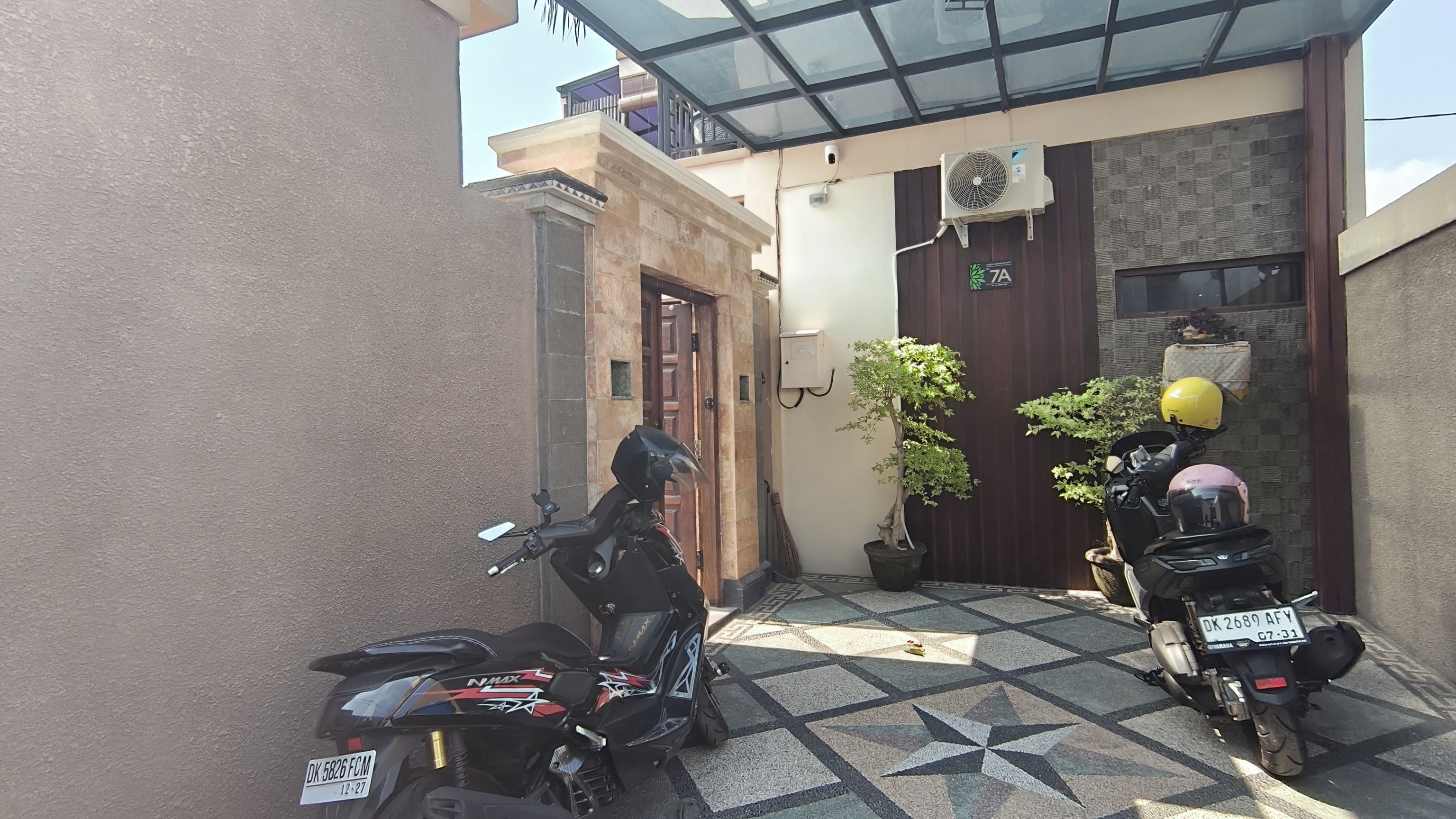 Une cour où sont garées deux motos, avec un sol carrelé, une porte en bois et une petite plante en pot.