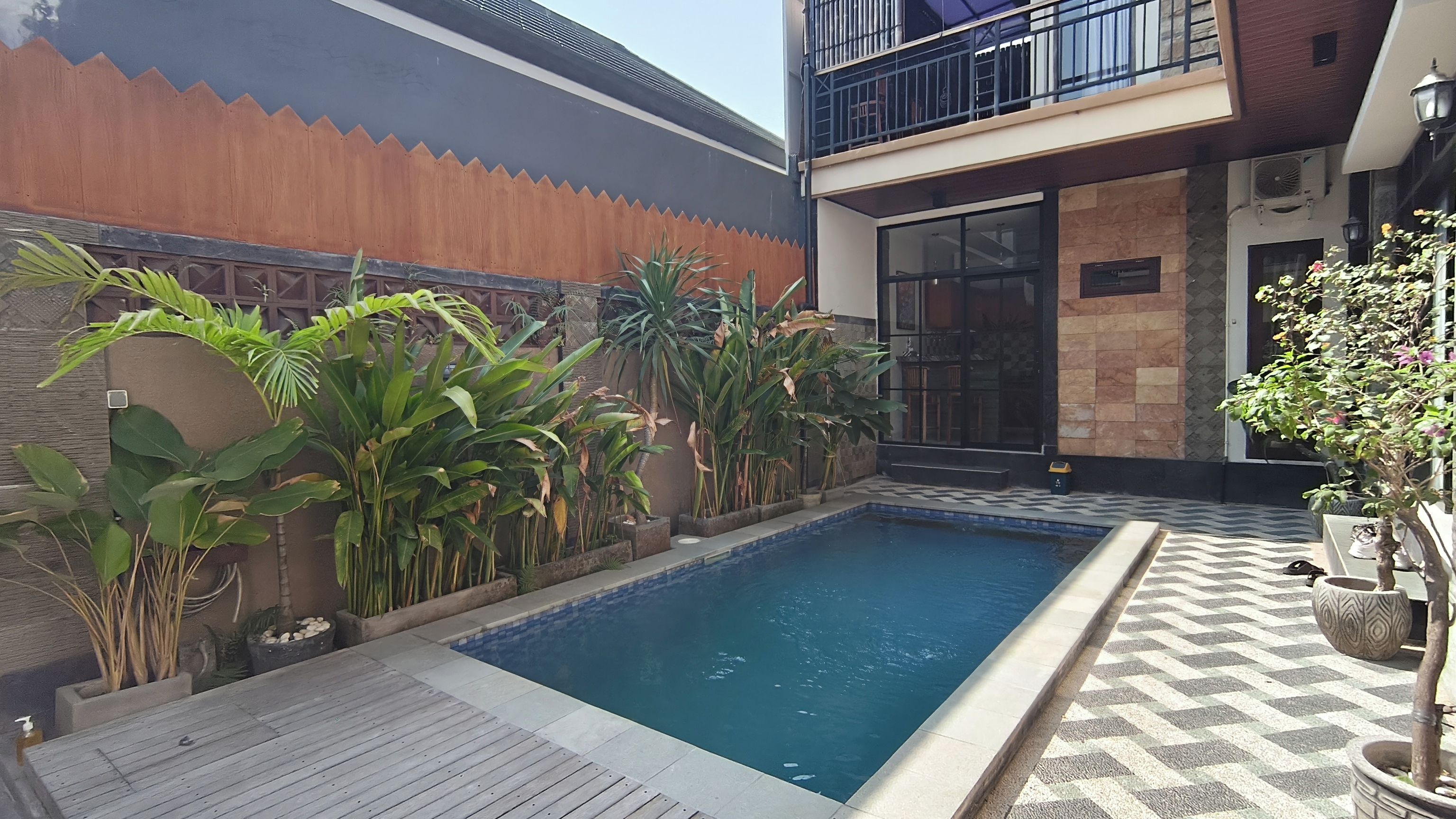 Une piscine dotée d’une terrasse en bois, entourée de plantes en pot et bordée d’un bâtiment moderne avec un balcon.