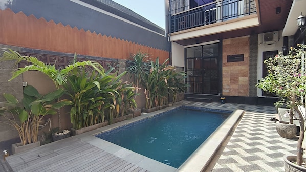 Une piscine dotée d’une terrasse en bois, entourée de plantes en pot et bordée d’un bâtiment moderne avec un balcon.