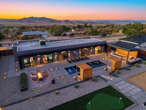A modern house with a swimming pool, outdoor seating, and a view of the mountains. - Pool + Spa + Putting Green + Bocce + Fire Pit | Natl Park West Gate | Desert Ace (Joshua Tree)
