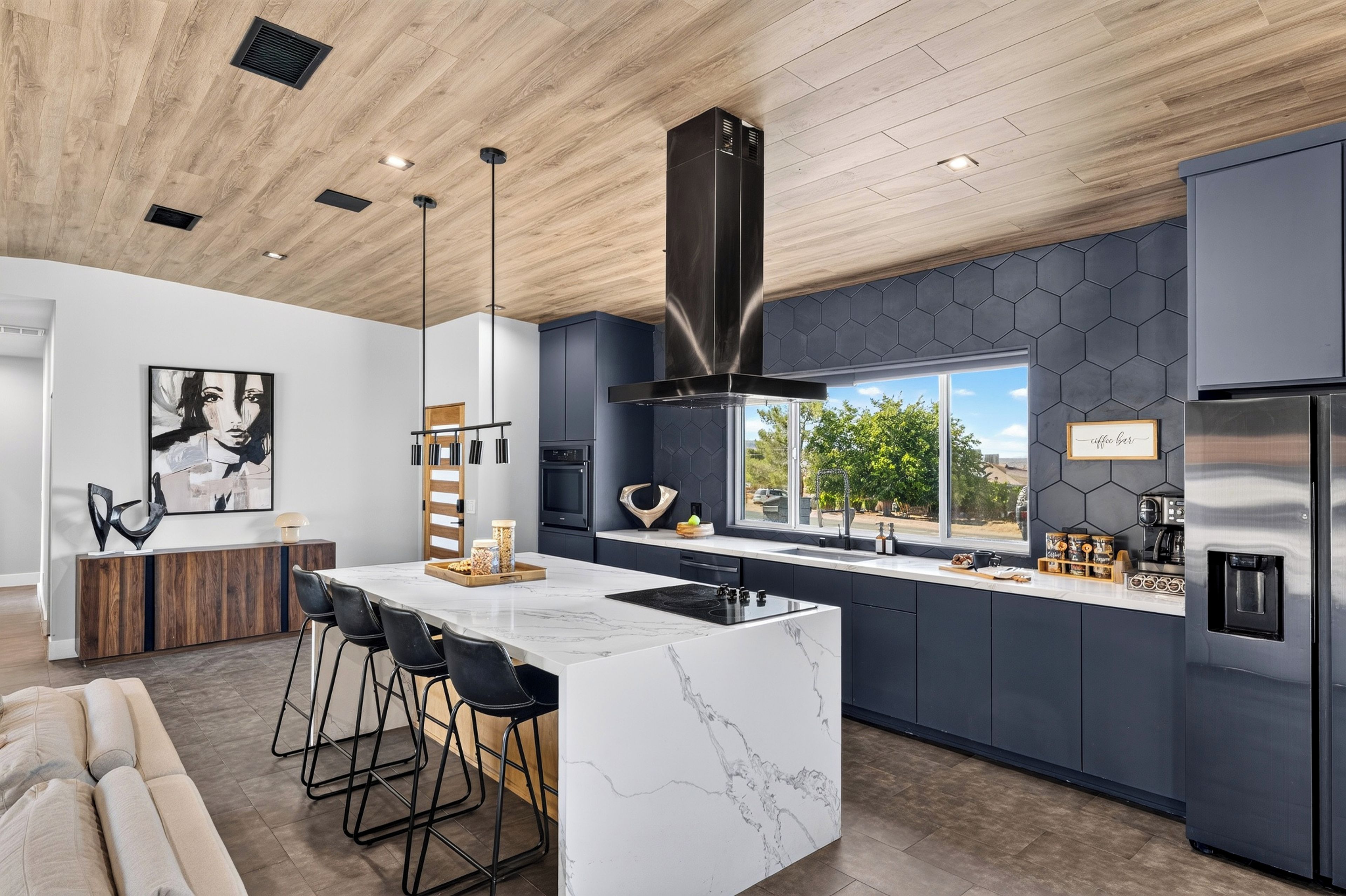 A modern kitchen with a marble island, bar stools, a large window, and a stainless steel refrigerator.