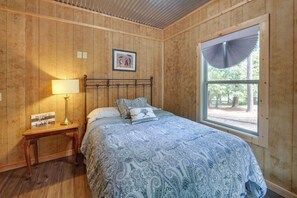 Une chambre avec des lambris en bois, un lit avec une literie à motifs, une table de chevet avec une lampe, un tableau encadré et une fenêtre donnant sur des arbres.