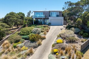 A modern two-story house with a driveway leading to a garage, surrounded by landscaped gardens and trees. - Stanley House Mt Martha. (Mount Martha)
