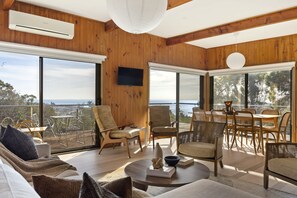 A spacious living area with a large window, a flat-screen TV mounted on the wall, and a dining area with wicker furniture. - Views at Yaringa McCrae (McCrae)