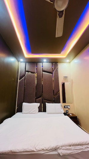 A hotel room with a bed, a bedside table, a nightstand, a mirror, and a ceiling fan. - Uday Hotel (Maura)