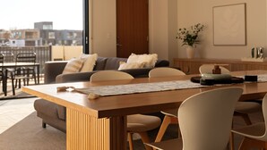 Una sala de estar moderna con comedor, que incluye una mesa de madera, sillas, un sofá y vistas de la ciudad a través de amplias ventanas.