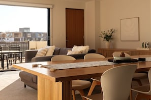 A modern living room with a dining area, featuring a wooden table, chairs, a sofa, and a view of the cityscape through large windows. - Waterfront Escape Rooftop Marina Views & Parking (Port Adelaide)
