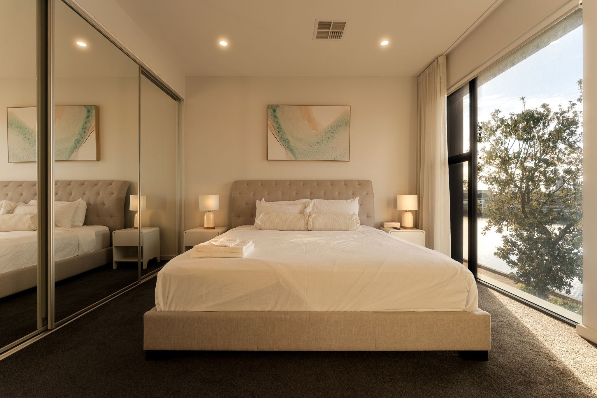 A modern bedroom with a large bed, two bedside tables, a mirror, and a view of trees through a window.