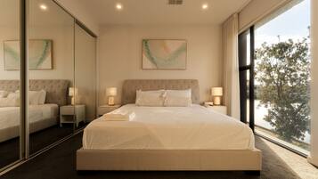 A modern bedroom with a large bed, two bedside tables, a mirror, and a view of trees through a window.