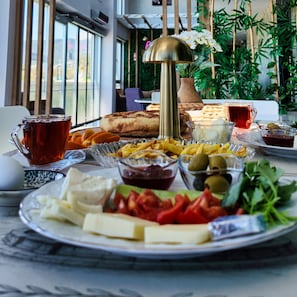 Ein Tisch reichhaltig mit Speisen gedeckt, darunter Brot, Käse, Oliven und Gemüse, mit einer Lampe und einer Vase im Hintergrund.
