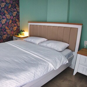 Una cama bien hecha con una colcha a rayas y almohadas, una mesita de noche con lámpara, y una cortina estampada en la pared.
