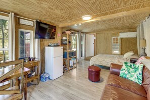 Ein gemütliches Blockhaus-Interieur mit Holzbalkendecke, einem an der Wand montierten Fernseher, einem Bett mit gemustertem Überwurf, einem braunen Ledersofa und einem Essbereich mit Holztisch und Stühlen.