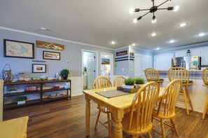 A dining area with a wooden table and chairs, a kitchen in the background, and a ceiling light fixture.